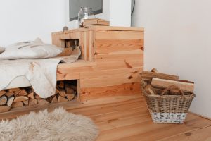 Przytulne wnętrze z łóżkiem z litego drewna wykonanym przez producenta łóżek drewnianych, z widocznym miejscem na opał i koszem z drewnem kominkowym.
