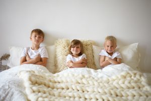 Trójka dzieci siedząca w łóżku, uśmiechnięta i gotowa do zabawy, symbolizująca radość wspólnego snu w łóżeczku piętrowym dla dzieci.