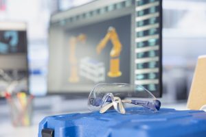 Roboty przemysłowe programowanie Ostrów Wielkopolski - widok na stanowisko pracy z monitorem wyświetlającym model robota oraz goglami ochronnymi na pierwszym planie.